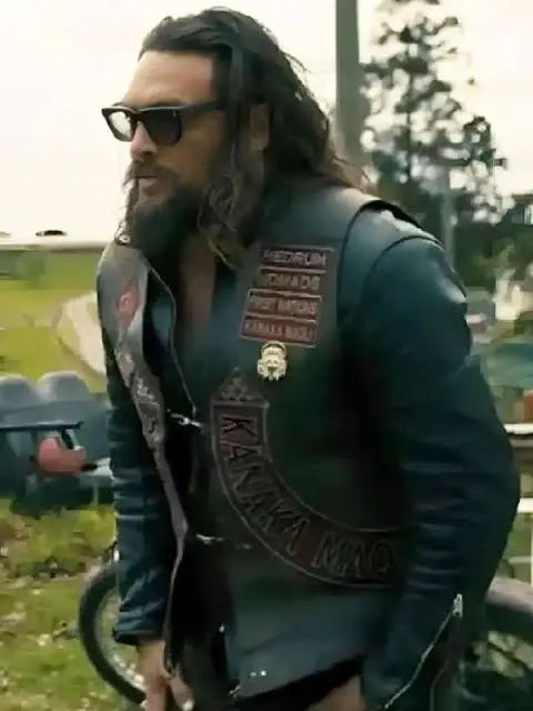 A man with long dark hair, a beard, and sunglasses wearing a black leather motorcycle vest with embroidered patches, standing outdoors near a motorcycle.