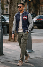 Man wearing a navy bomber jacket with a Captain America logo, gray sweater, beige pants, and brown shoes on a city street.