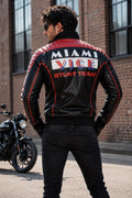 A man wearing a black leather jacket with red piping and a large 'MIAMI VICE STUNT TEAM' graphic on the back, standing outdoors in front of a brick building with a motorcycle visible on the left.