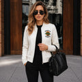 Woman wearing a white bomber jacket with a logo, black pants, and sunglasses, holding a black handbag on a city street.