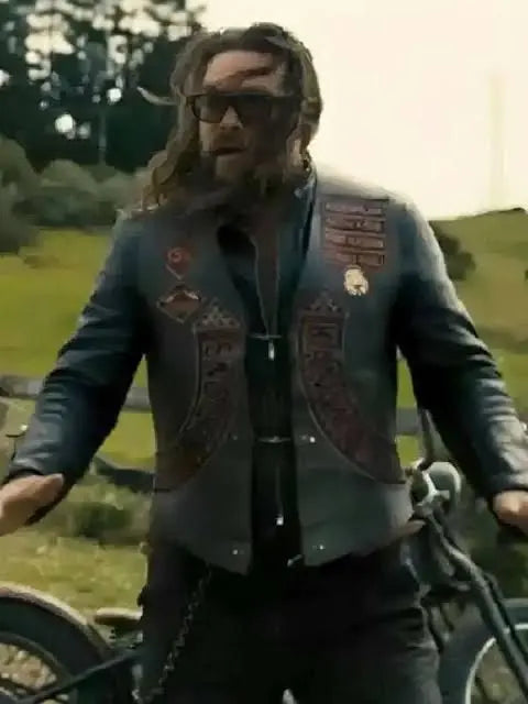 A man with a beard and sunglasses wearing a dark leather motorcycle vest with decorative patches, standing outdoors next to a motorcycle.